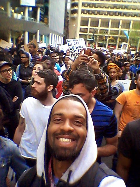 A group of diverse protesters participating in a march, with one person in the foreground smiling at the camera. Signs are held up in the background, with visible phrases supporting freedom.